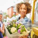 A young woman stands at the front door of her home and reads the card from a big bouquet of flowers . Behind her is the delivery girl who has just dropped them off. The woman looks proudly to camera whilst holding her floral surprise.